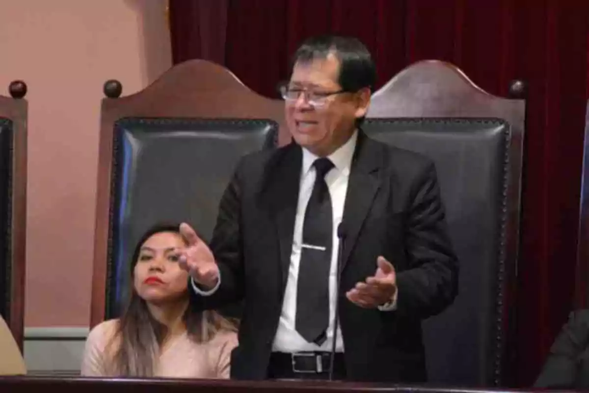 Hombre de traje negro hablando de pie frente a un micrófono en una sala con sillas grandes y fondo de cortinas rojas Hombre de traje negro hablando de pie frente a un micrófono en una sala con sillas grandes y fondo de cortinas rojas