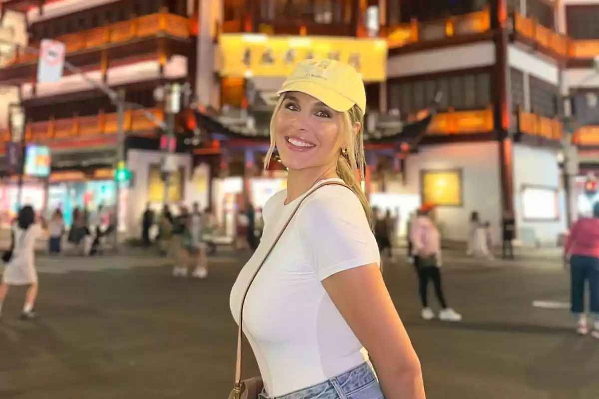 Una persona sonriente con gorra y camiseta blanca posa en una calle iluminada con edificios de estilo tradicional al fondo.