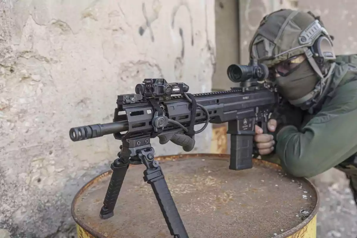 Un soldado con equipo táctico apunta con un rifle de asalto apoyado en un barril oxidado.