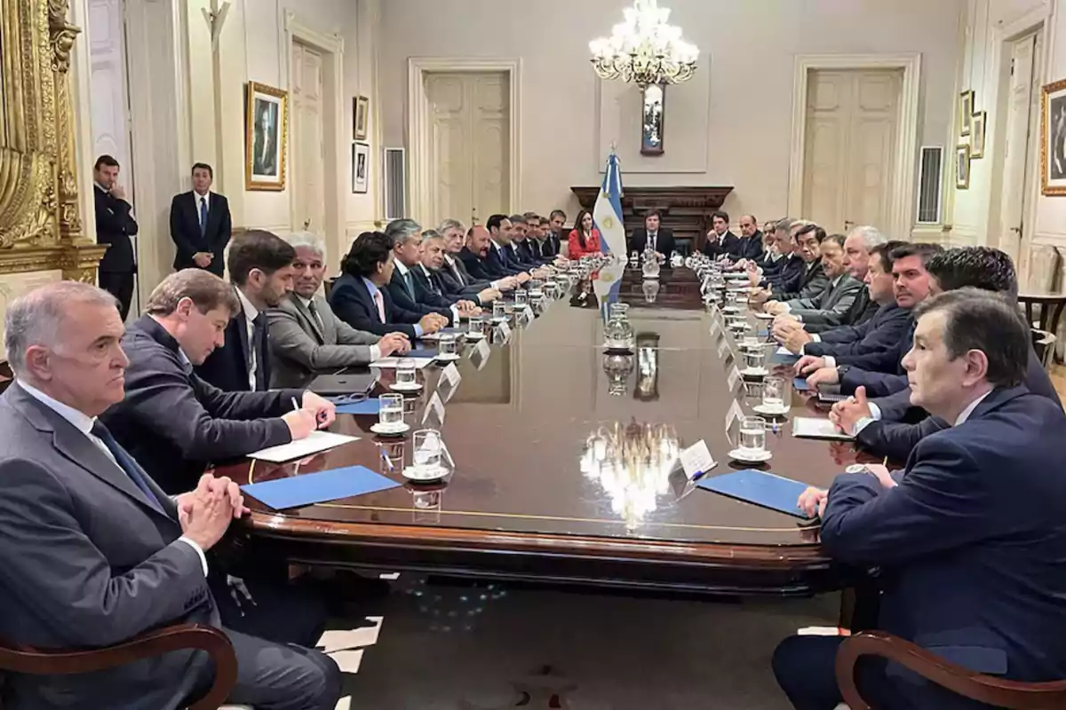 A group of people is gathered around a large table in an elegant room with an Argentine flag in the background.