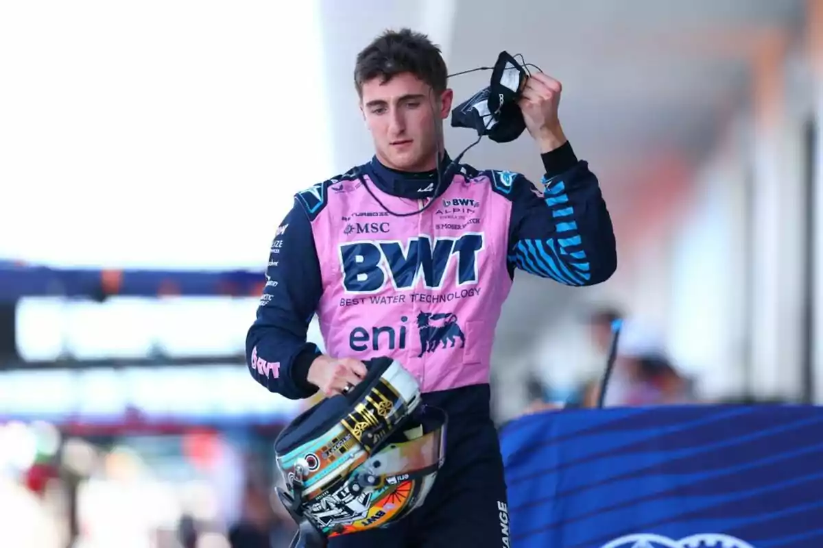 A race car driver in a pink and blue suit holds a helmet and a mask while walking down the pit lane. A race car driver in a pink and blue suit holds a helmet and a mask while walking down the pit lane.
