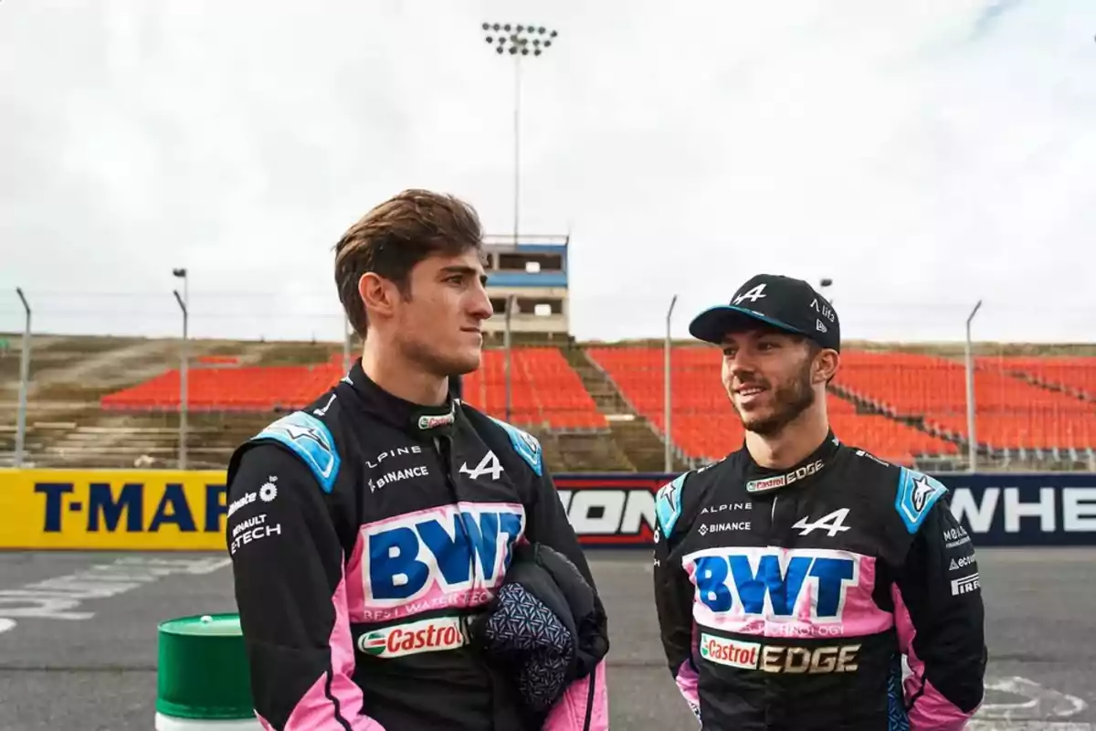 Two racing drivers in Alpine team racing suits talk on a track with empty stands in the background. Two racing drivers in Alpine team racing suits talk on a track with empty stands in the background.