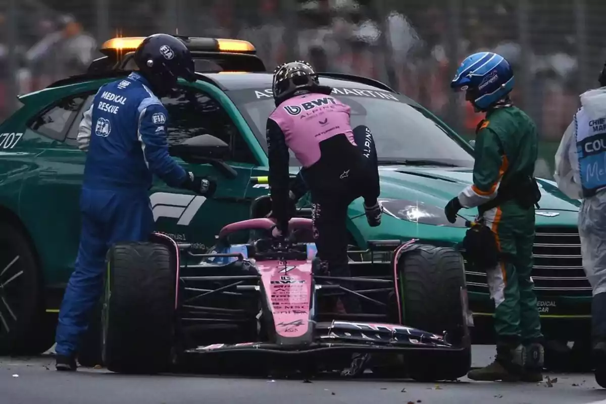 A race car driver in a pink and blue suit is getting out of a Formula 1 car while being assisted by medical rescue personnel dressed in blue and green, with a green safety vehicle in the background. A race car driver in a pink and blue suit is getting out of a Formula 1 car while being assisted by medical rescue personnel dressed in blue and green, with a green safety vehicle in the background.