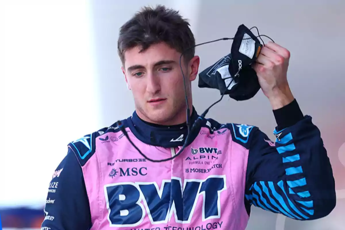 A race car driver in a pink and blue suit holds a black garment in his hand.