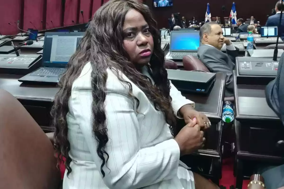 Una persona sentada en un salón con computadoras portátiles y banderas en el fondo.