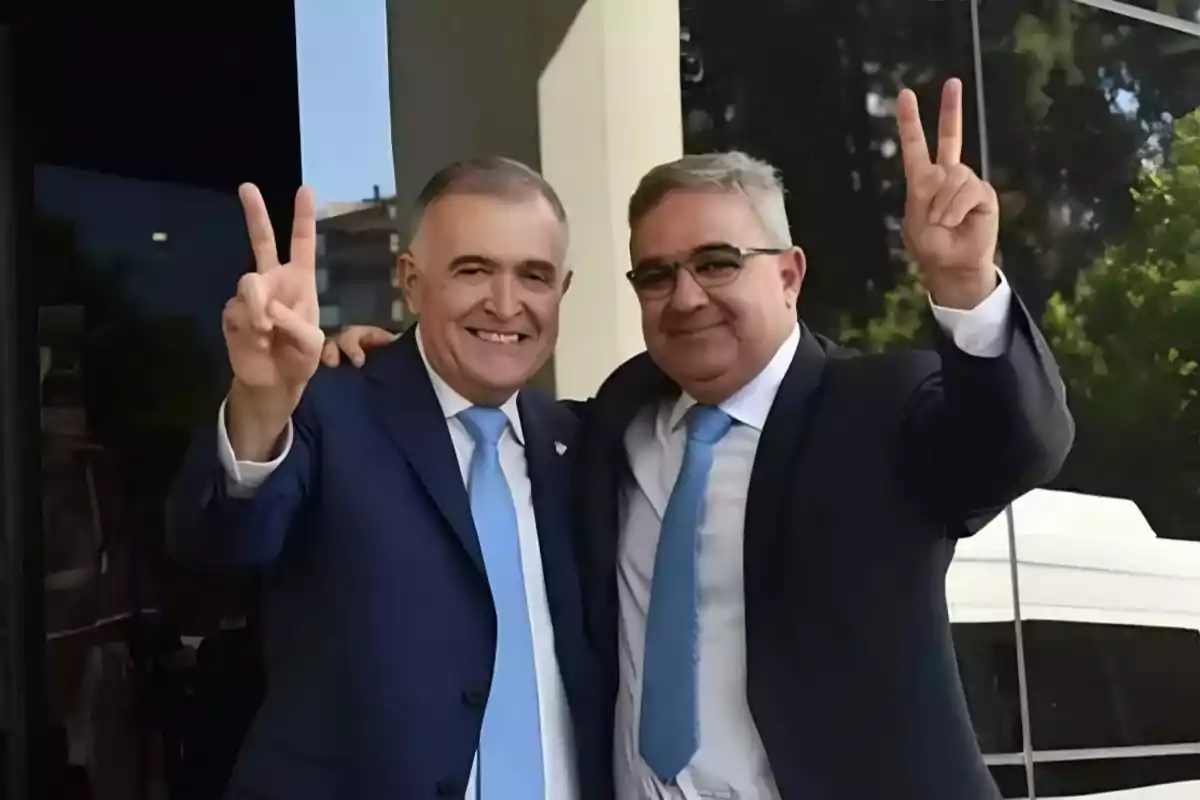 Dos hombres sonrientes con trajes y corbatas azules levantan las manos haciendo el signo de la paz.