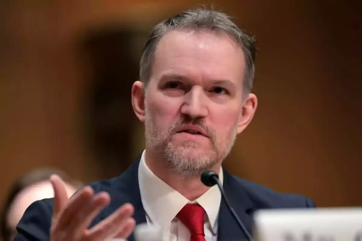 Middle-aged man with a beard and short hair wearing a suit and red tie speaking in front of a microphone in a formal setting Middle-aged man with a beard and short hair wearing a suit and red tie speaking in front of a microphone in a formal setting