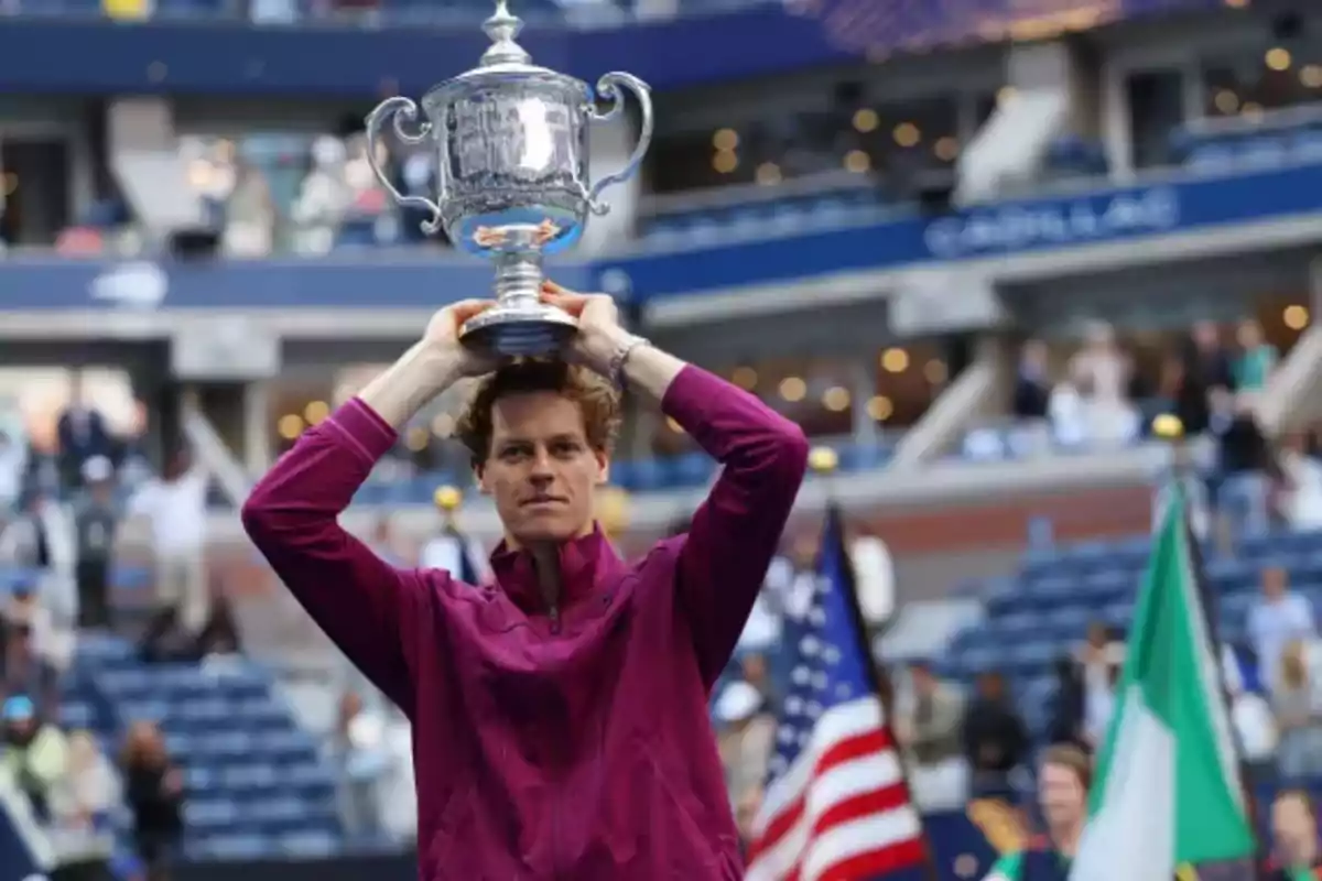 Persona sosteniendo un trofeo plateado sobre su cabeza en un estadio de tenis.