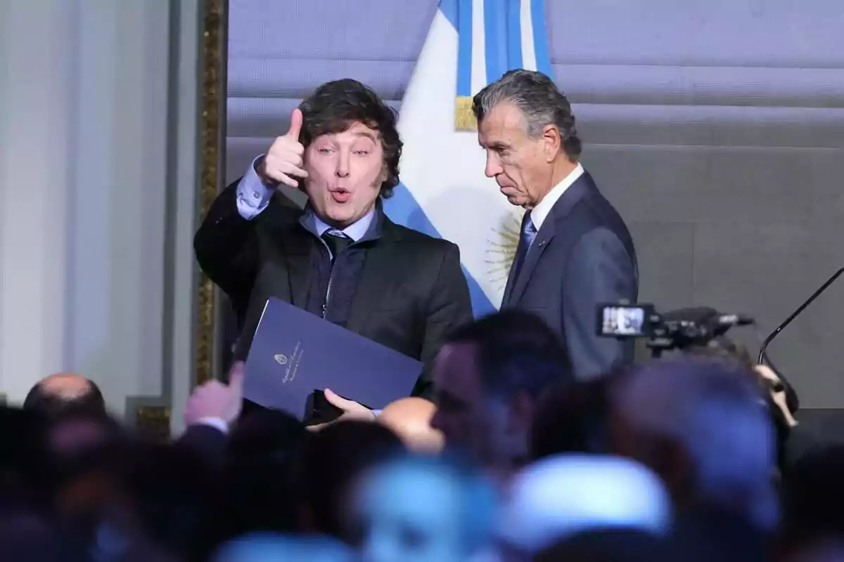 Dos hombres en un evento formal con una bandera de Argentina de fondo uno de ellos gesticula con la mano mientras sostiene un documento.