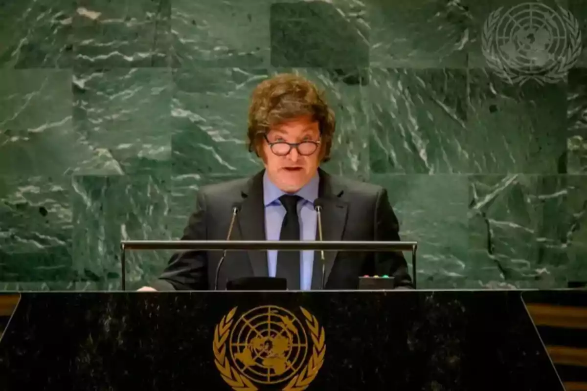 A man with glasses and a dark suit speaks from a podium with the United Nations emblem on a green marble background.