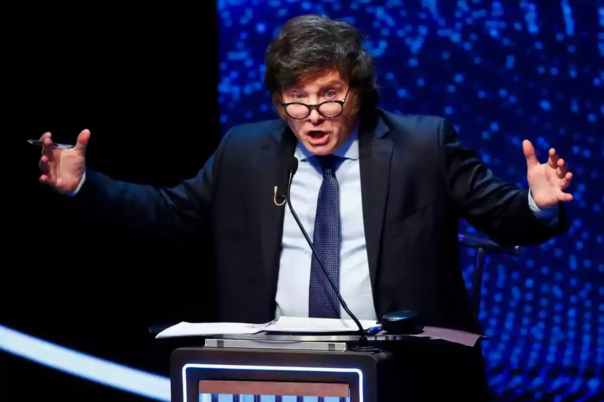 Un hombre con gafas y traje oscuro gesticula mientras habla en un podio con un fondo azul.