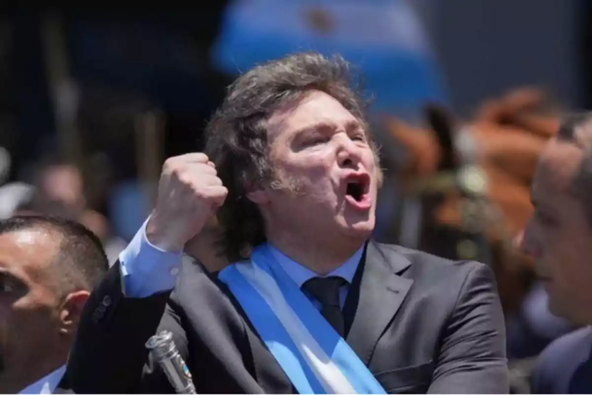 A man in a suit and a blue and white sash raises his fist as he appears to be speaking passionately at an outdoor event.