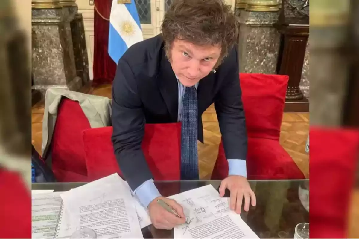 Un hombre de traje y corbata está firmando documentos en una mesa con una bandera de Argentina al fondo.