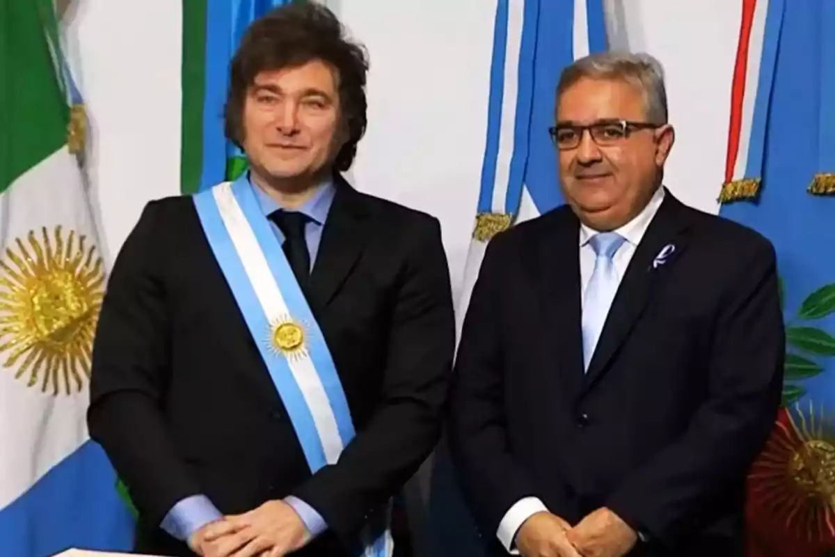 Dos hombres de pie con trajes formales y bandas presidenciales, frente a banderas argentinas.