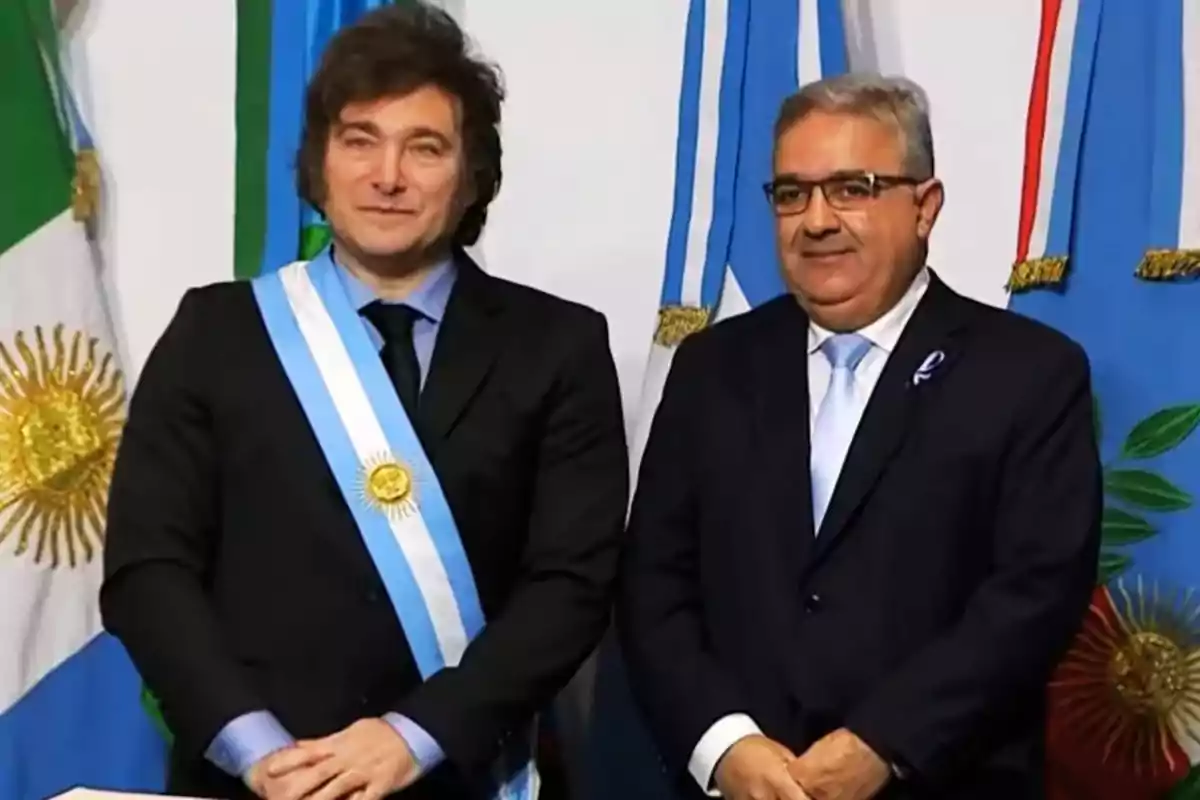 Dos hombres de pie con trajes oscuros y corbatas azules, uno de ellos lleva una banda presidencial, frente a banderas argentinas.