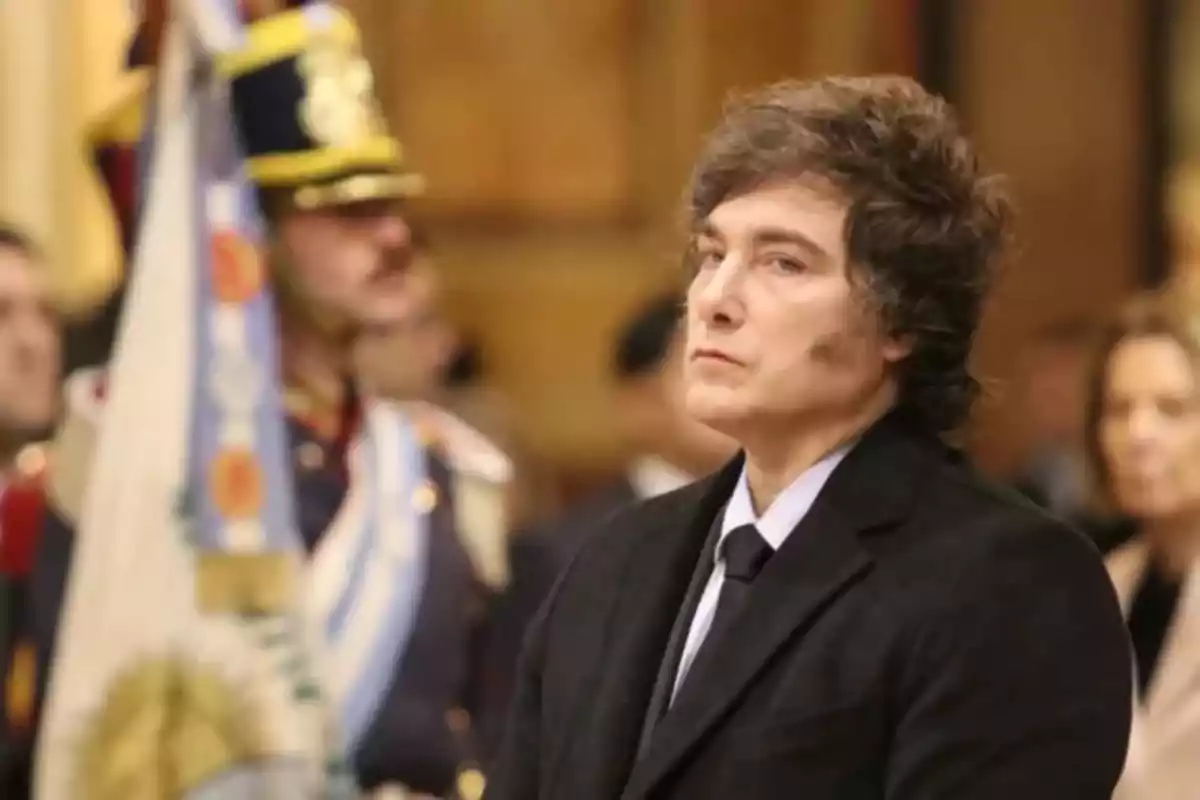 Wavy-haired man in a dark suit at a formal event with the Argentine flag and a uniformed guard in the background