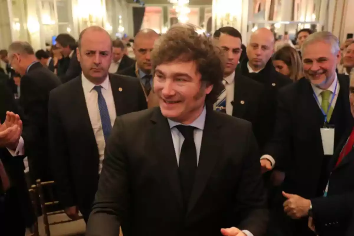 Un hombre sonriente con traje oscuro rodeado de personas en un evento formal.