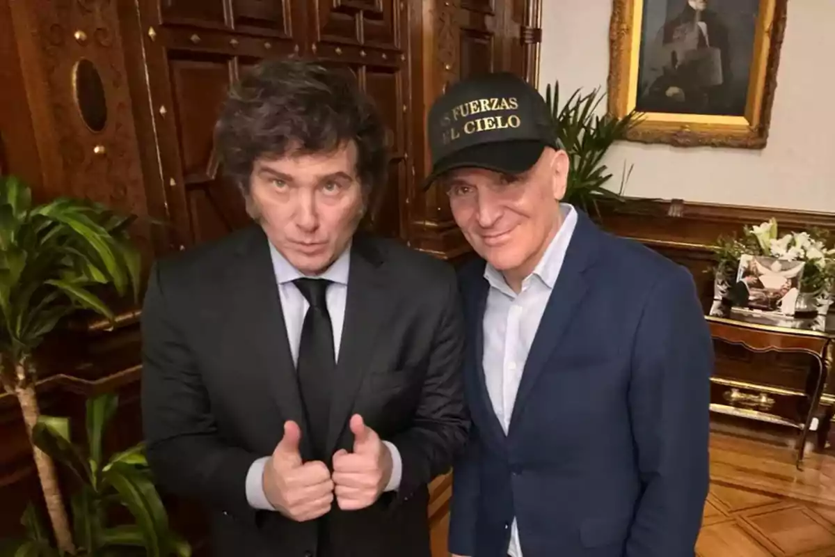 Dos hombres posan juntos en una oficina elegante, uno de ellos lleva traje y corbata y muestra ambos pulgares hacia arriba, el otro viste saco azul y gorra negra con letras amarillas