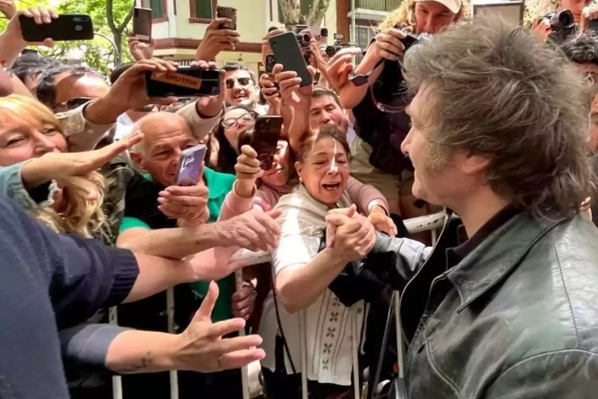 Una multitud de personas emocionadas rodea a un hombre que está saludando y estrechando la mano de una mujer mientras otros toman fotos con sus teléfonos móviles.