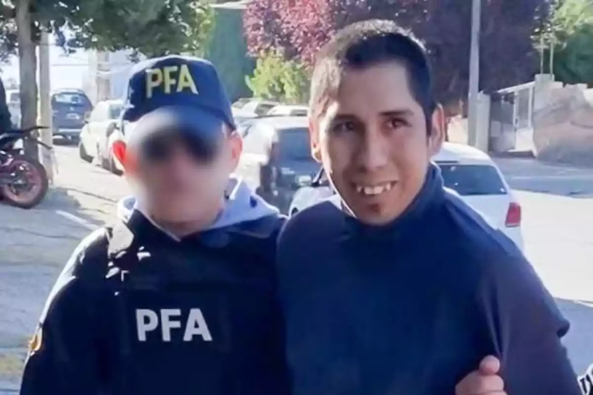 A man in a dark sweater is escorted by a PFA officer on a street with parked cars and trees in the background.