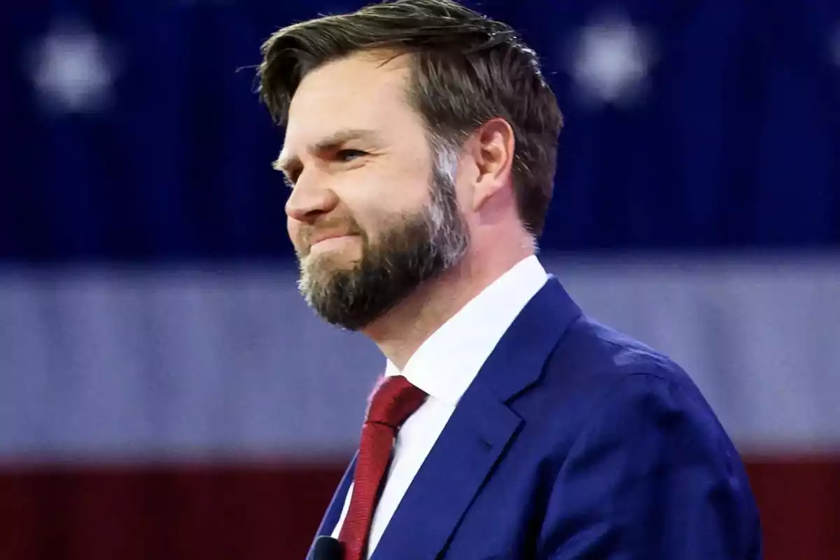 Hombre de barba y cabello corto con traje azul y corbata roja frente a una bandera de Estados Unidos desenfocada Hombre de barba y cabello corto con traje azul y corbata roja frente a una bandera de Estados Unidos desenfocada