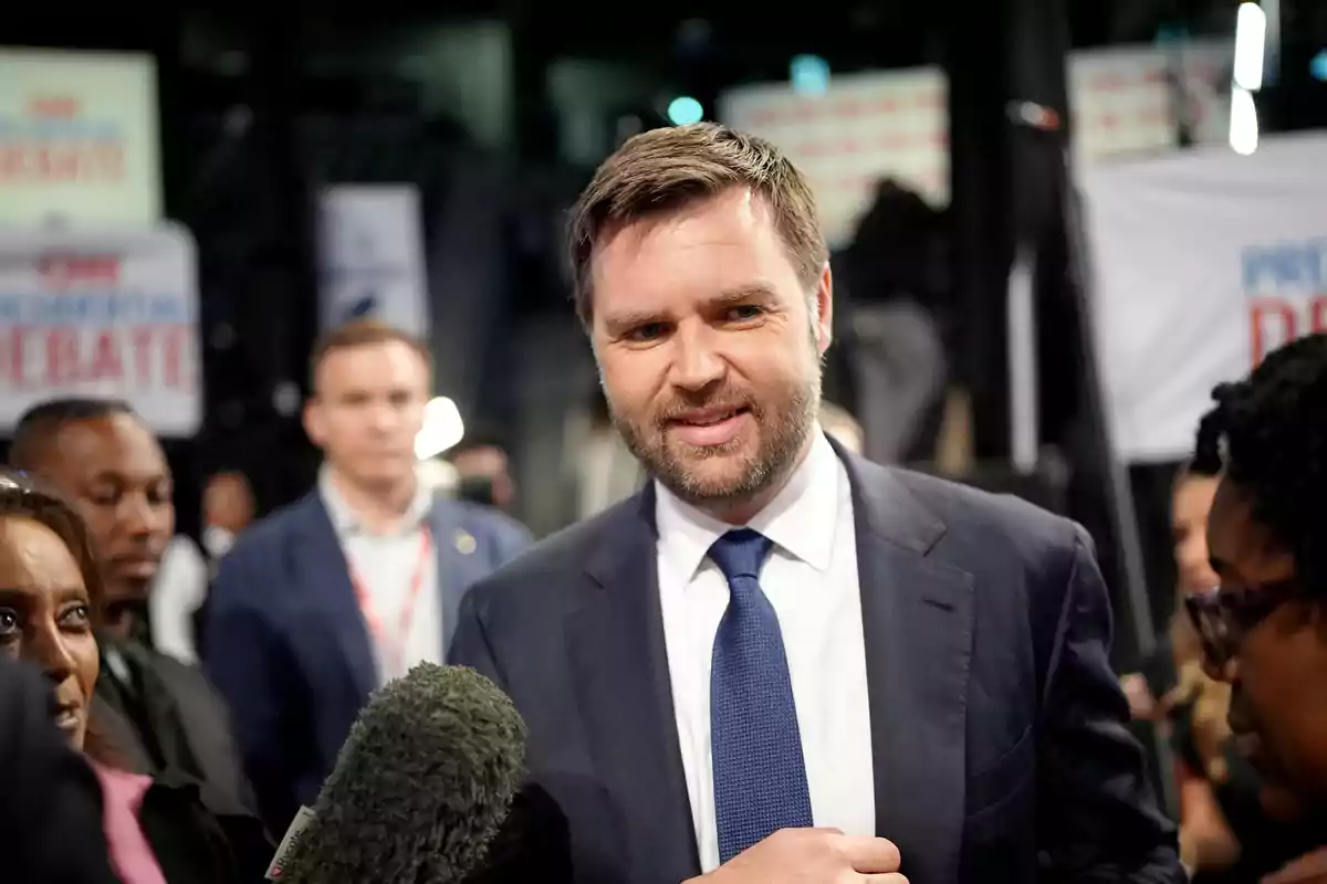Un hombre con traje y corbata azul está rodeado de personas y micrófonos en un evento, con carteles de debate al fondo. Un hombre con traje y corbata azul está rodeado de personas y micrófonos en un evento, con carteles de debate al fondo.
