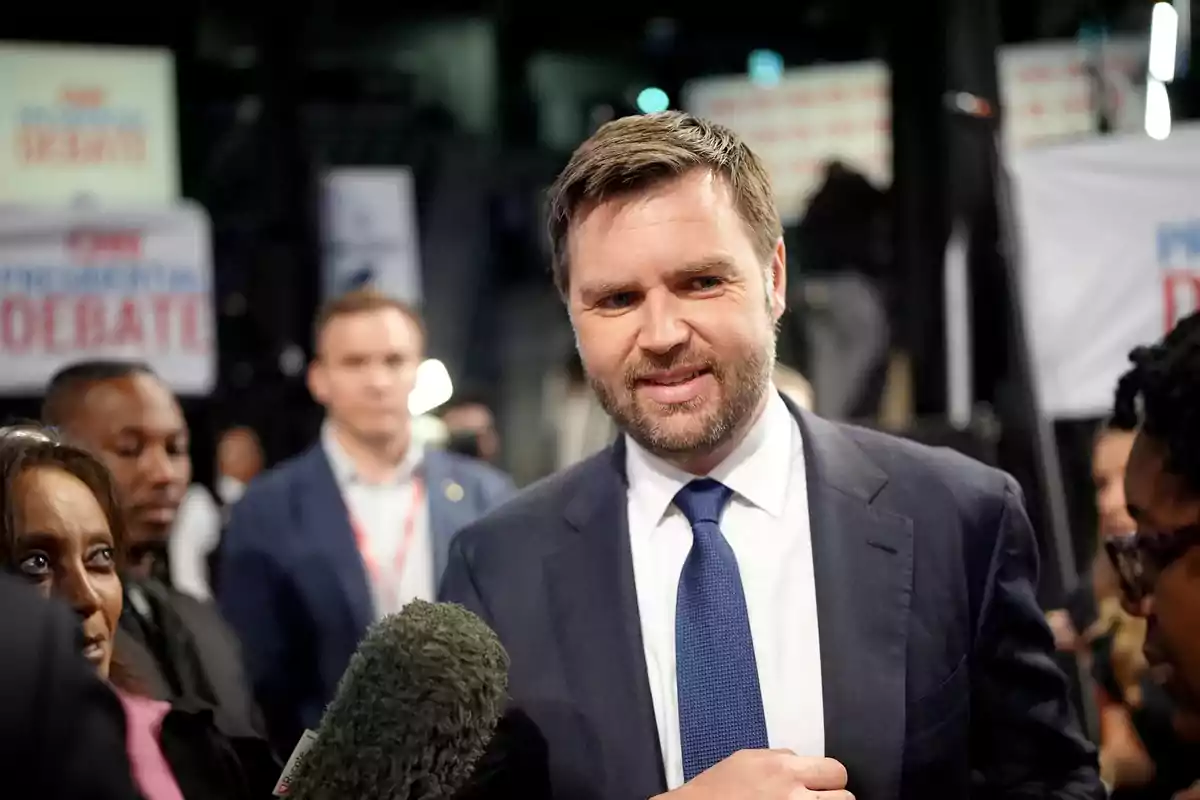 Hombre con traje y corbata azul siendo entrevistado por la prensa en un evento con carteles de debate al fondo Hombre con traje y corbata azul siendo entrevistado por la prensa en un evento con carteles de debate al fondo