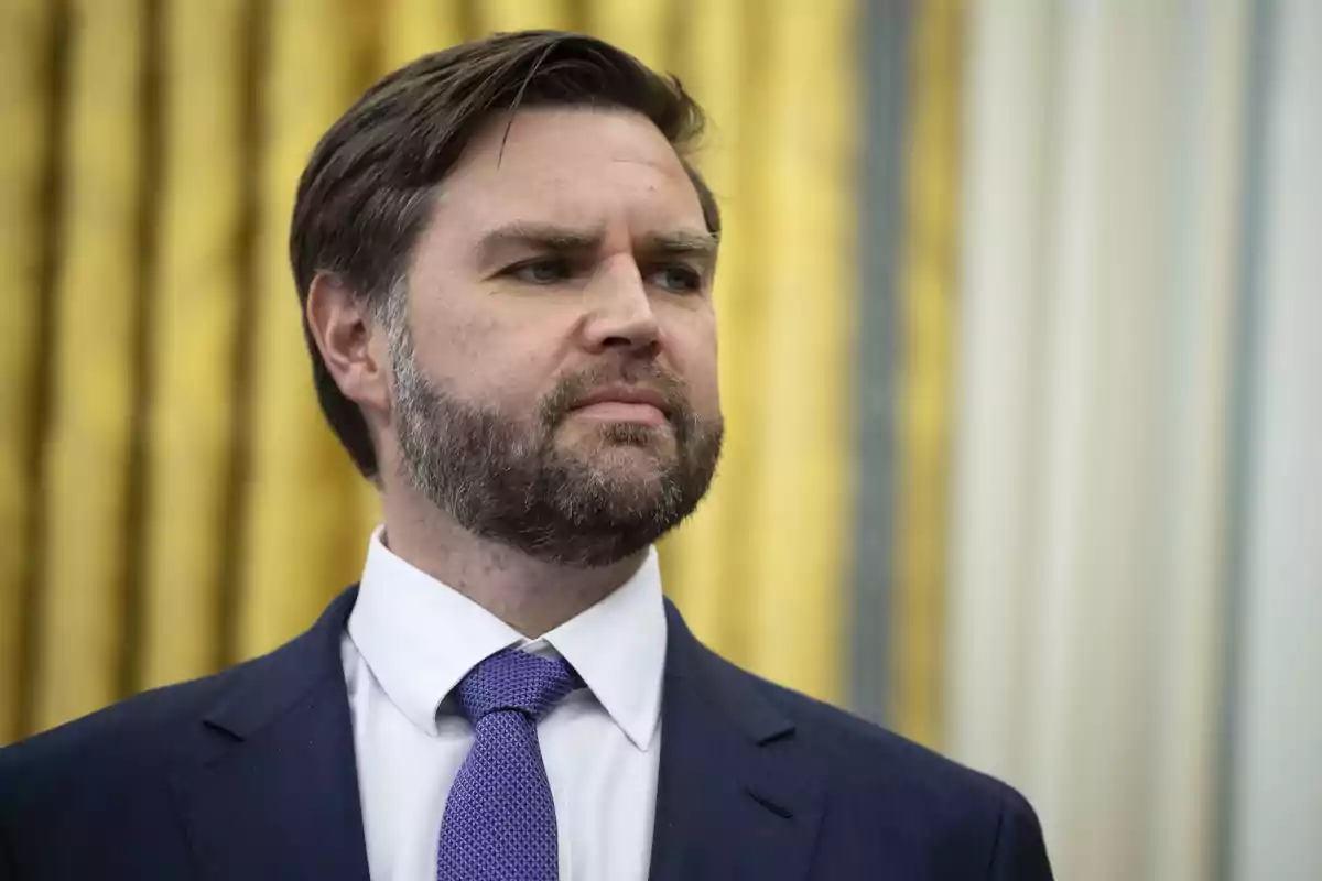 Un hombre con barba y traje azul mira hacia un lado con expresión seria frente a un fondo con cortinas doradas. Un hombre con barba y traje azul mira hacia un lado con expresión seria frente a un fondo con cortinas doradas.