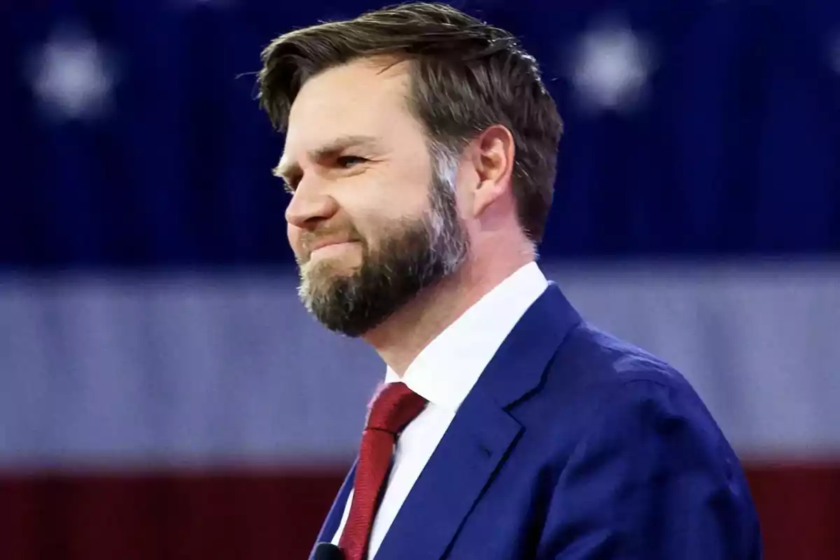 Man with a beard and short hair wearing a blue suit and red tie in front of a background with white stars on blue and red and white stripes