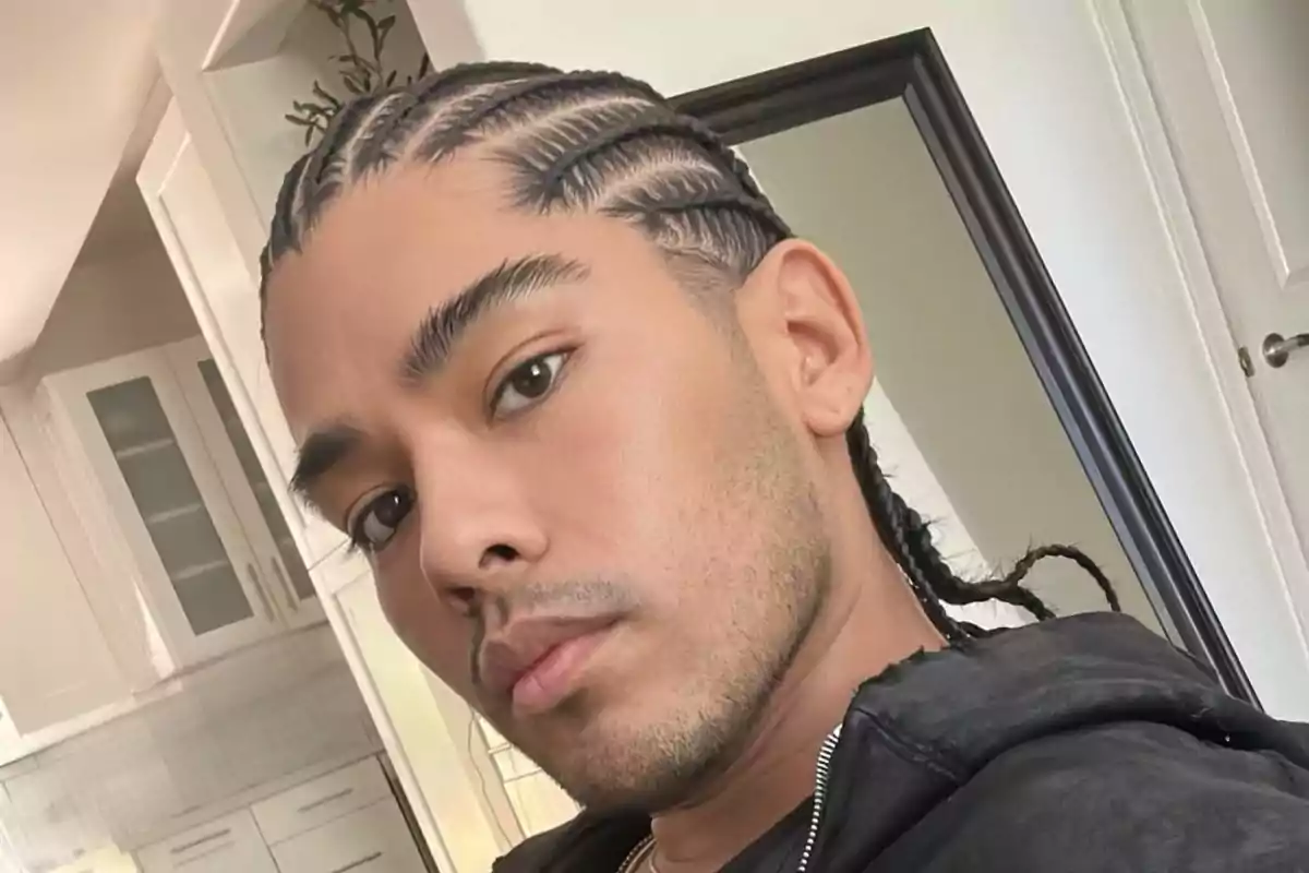 Un hombre con trenzas posando frente a un espejo en una habitación con gabinetes blancos.