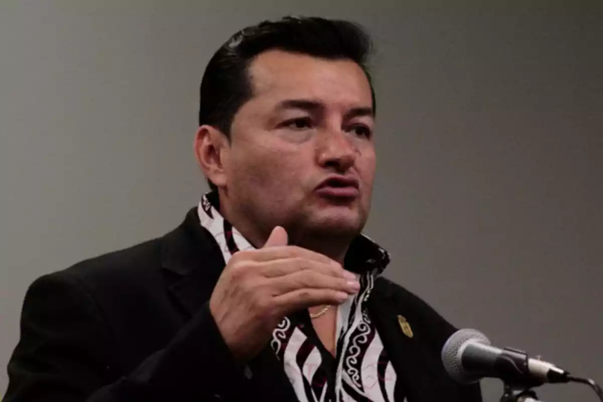 Hombre de cabello oscuro hablando frente a un micrófono con gesto serio y mano levantada