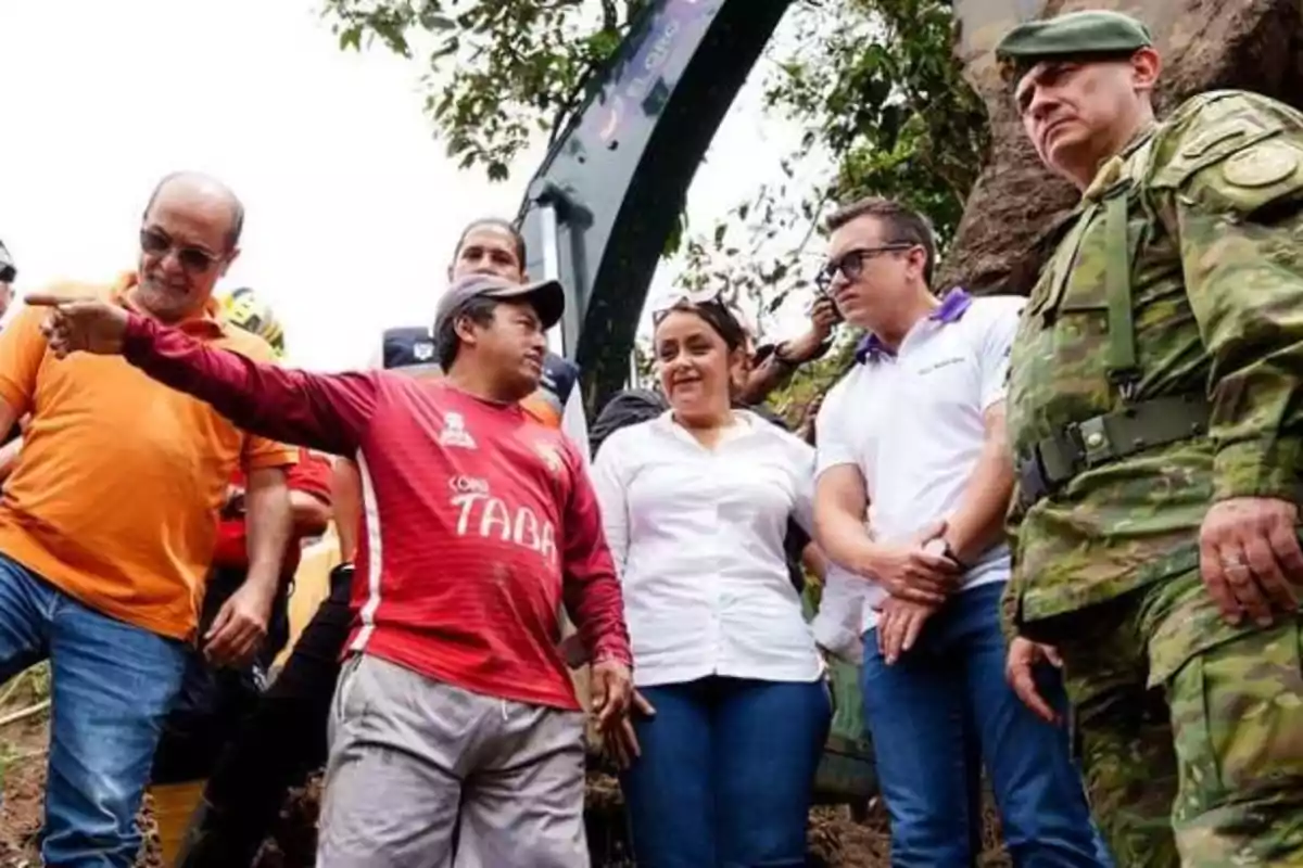 Un grupo de personas, incluyendo un hombre con uniforme militar, están de pie al aire libre, mientras uno de ellos señala algo en la distancia.