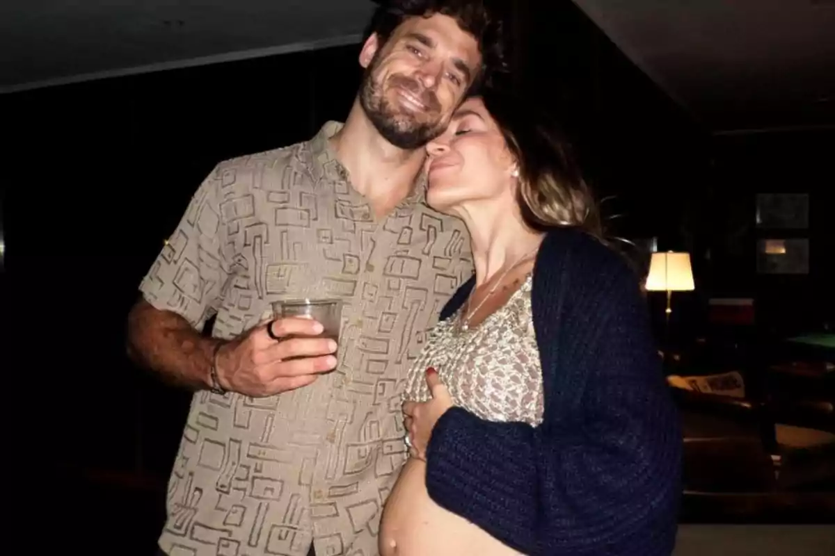 Couple hugging and smiling while the pregnant woman caresses her belly and the man holds a glass