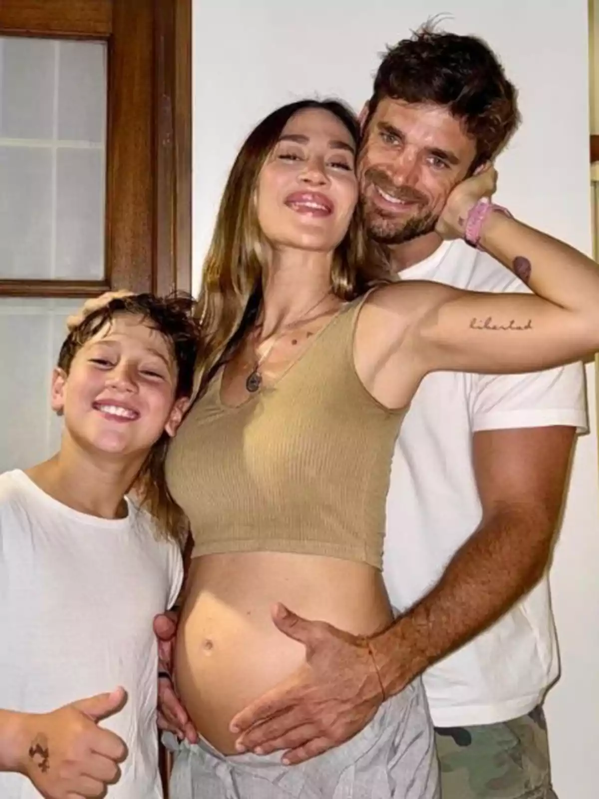 Familia sonriente posando junta una mujer embarazada en el centro con un hombre abrazándola por detrás y un niño a su lado mostrando el pulgar arriba Familia sonriente posando junta una mujer embarazada en el centro con un hombre abrazándola por detrás y un niño a su lado mostrando el pulgar arriba