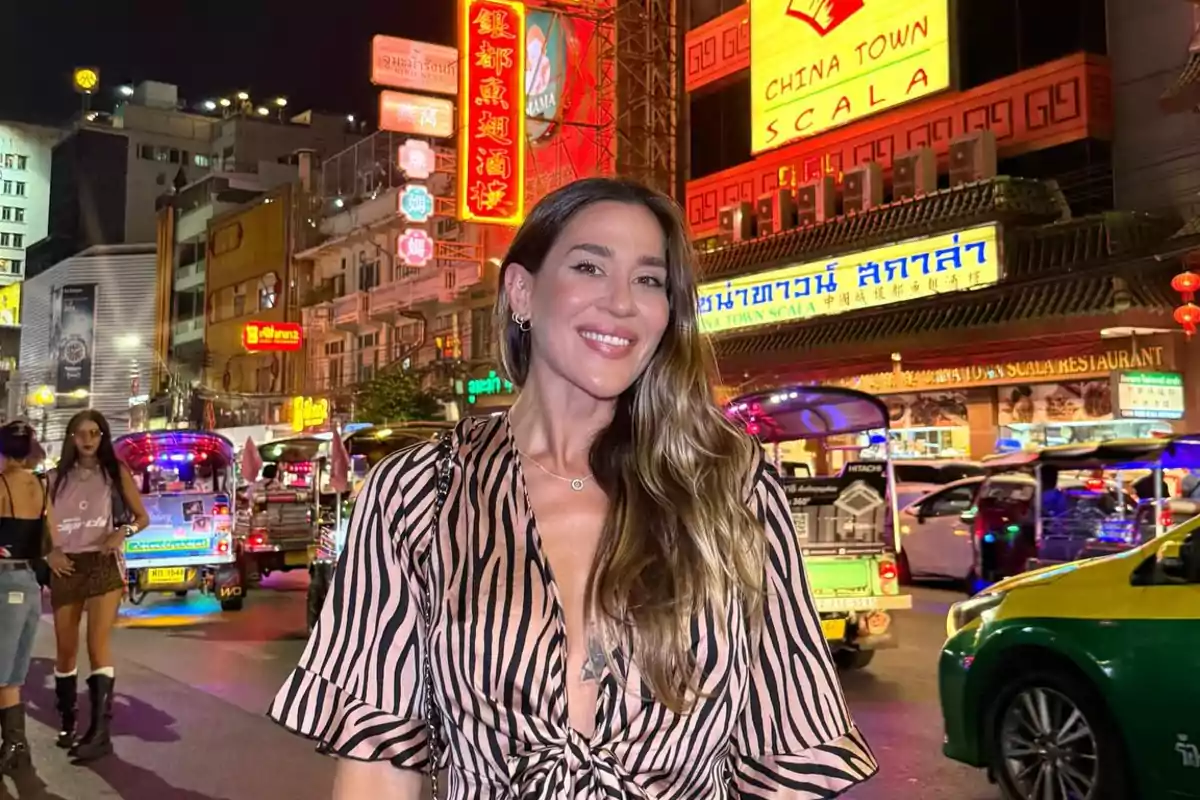 Una mujer sonriente posa en una calle iluminada de un barrio chino, rodeada de letreros de neón y tráfico nocturno.