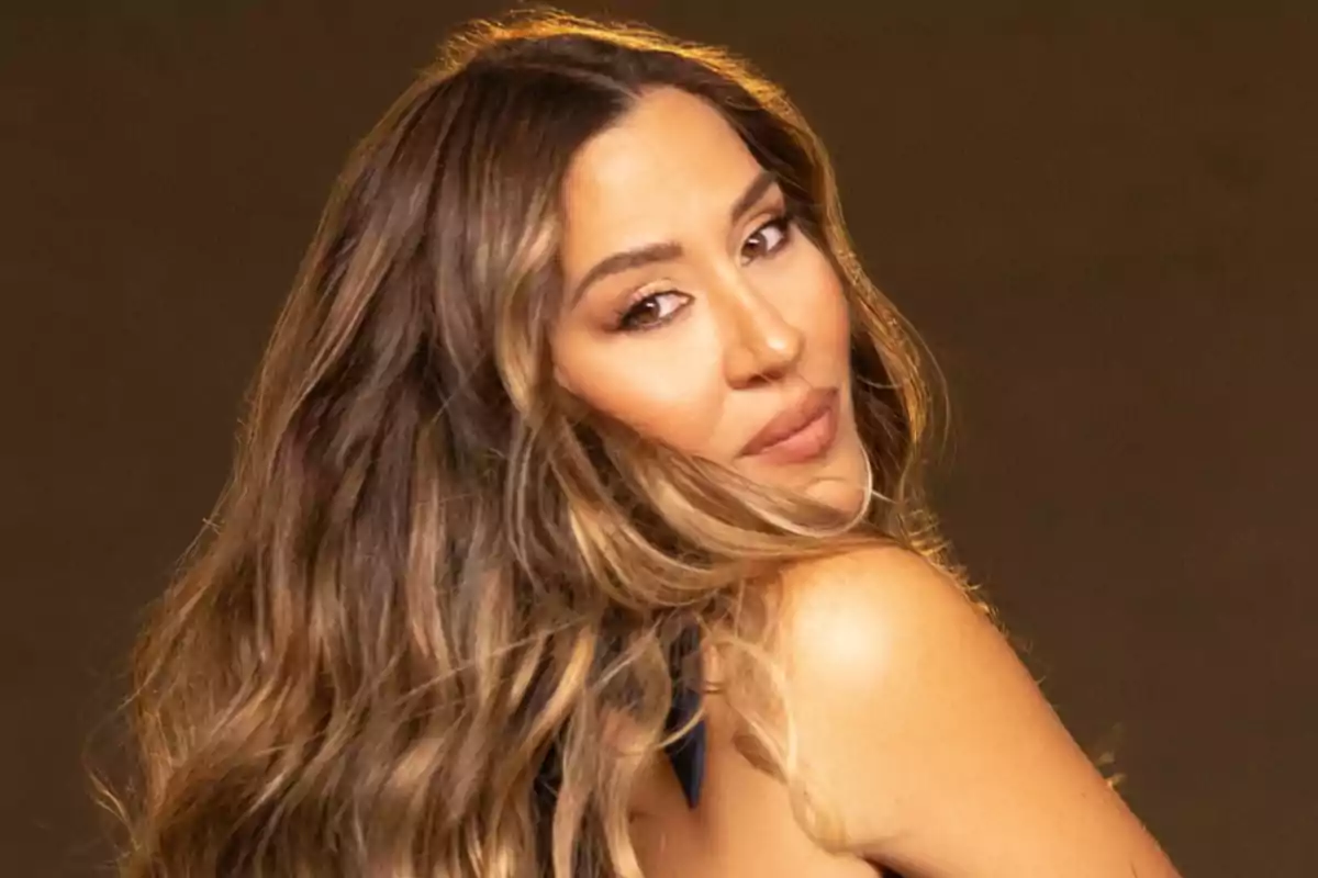 A person with long, wavy hair looking back with a gentle expression and a brown background.