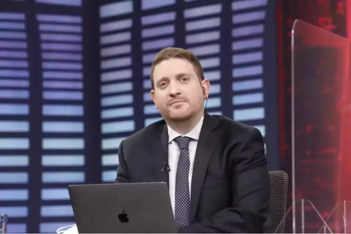 Un hombre con traje y corbata está sentado frente a una laptop en un estudio de televisión con un fondo de luces azules y rojas.