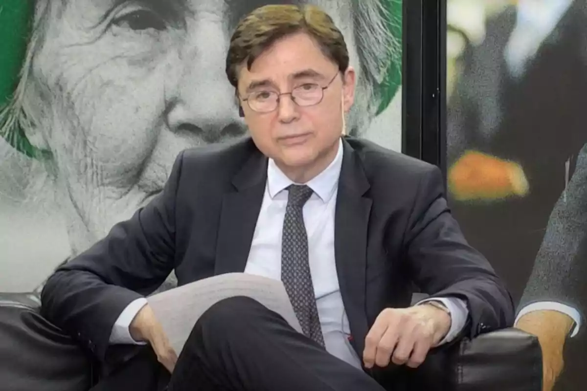 Un hombre con traje y corbata está sentado en un sillón, sosteniendo un papel, con un fondo que muestra una imagen en blanco y negro de una persona mayor. Un hombre con traje y corbata está sentado en un sillón, sosteniendo un papel, con un fondo que muestra una imagen en blanco y negro de una persona mayor.