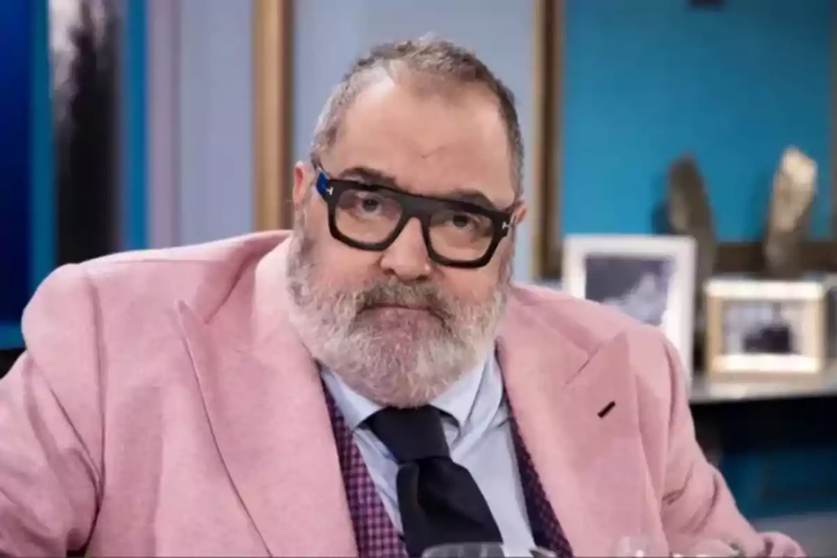 Un hombre con barba y gafas negras lleva un traje rosa y corbata oscura en un entorno interior.