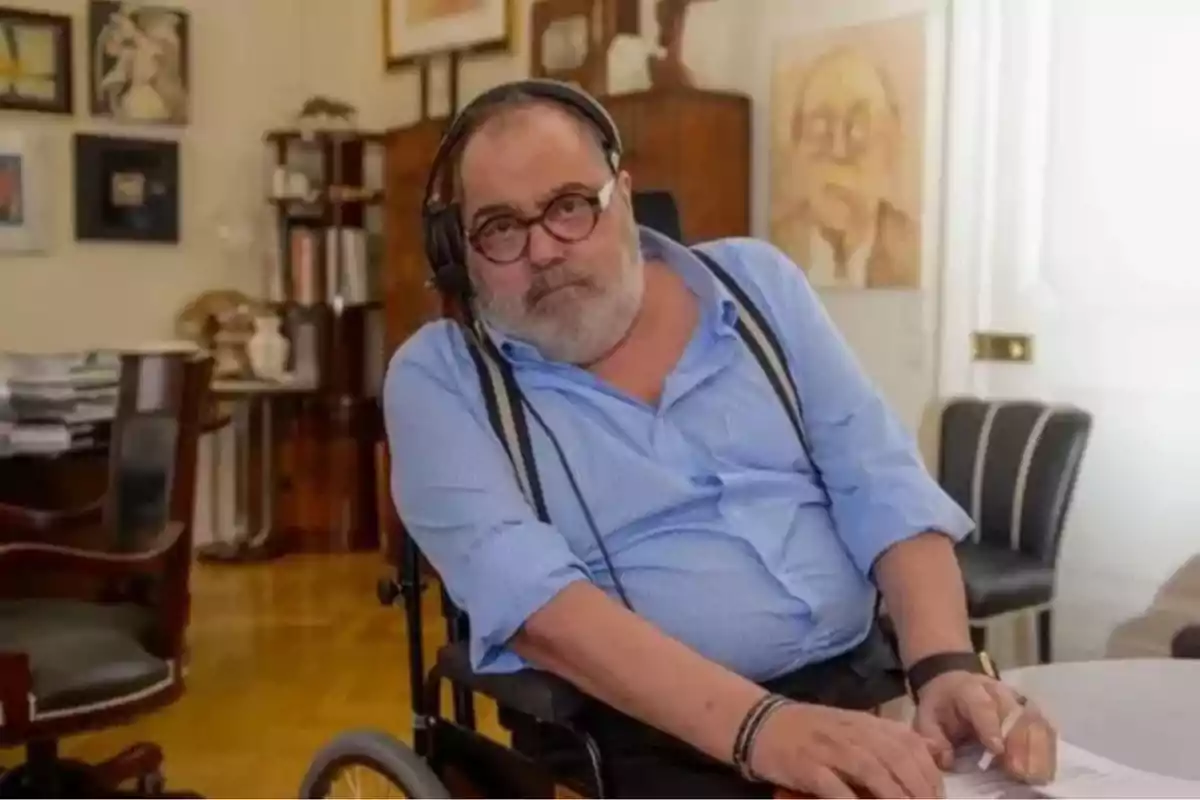 Un hombre con barba y gafas está sentado en una silla de ruedas en una oficina rodeada de arte y libros.