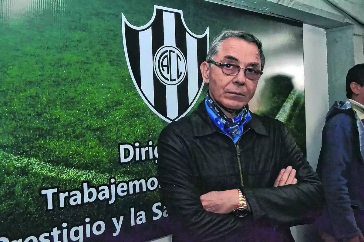 Gray-haired man with glasses, arms crossed, standing in front of a soccer crest on a wall with grass in the background and motivational text. Gray-haired man with glasses, arms crossed, standing in front of a soccer crest on a wall with grass in the background and motivational text.