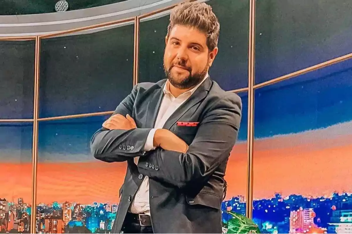 Un hombre con traje oscuro y camisa blanca, con los brazos cruzados, de pie frente a un fondo que muestra un paisaje urbano al atardecer. Un hombre con traje oscuro y camisa blanca, con los brazos cruzados, de pie frente a un fondo que muestra un paisaje urbano al atardecer.