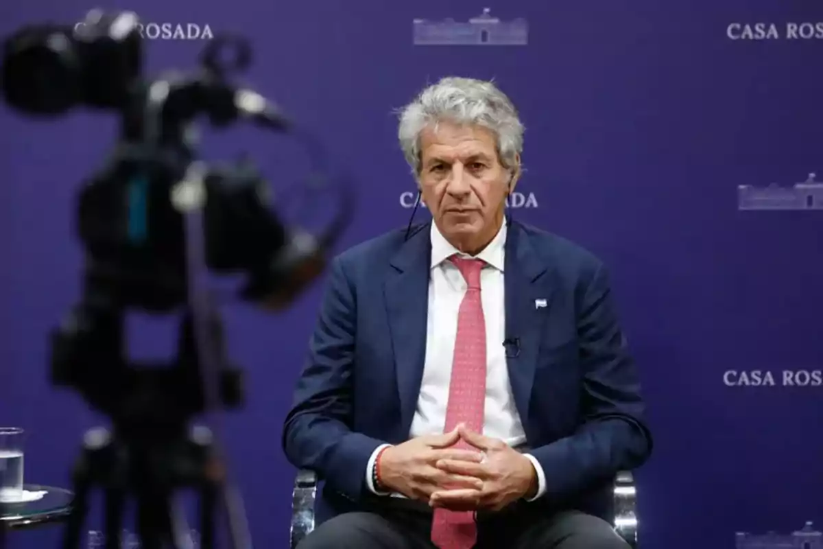Gray-haired man in a suit and tie sitting in front of a camera on a set with a purple background that says Casa Rosada Gray-haired man in a suit and tie sitting in front of a camera on a set with a purple background that says Casa Rosada