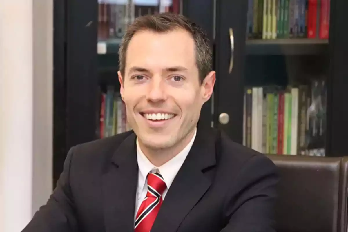 Un hombre sonriendo con traje y corbata roja sentado frente a un estante de libros.