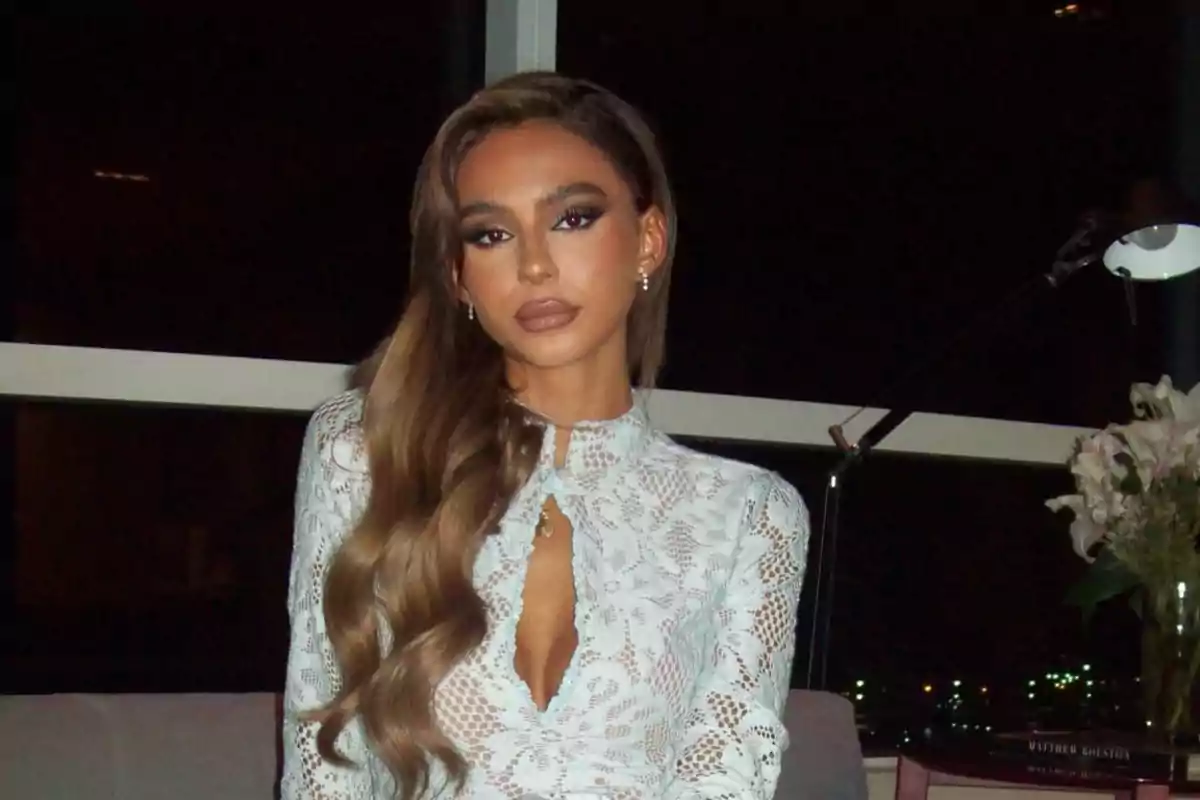 Una persona con cabello largo y ondulado lleva un vestido de encaje blanco sentada en un sofá con una lámpara y flores al fondo.