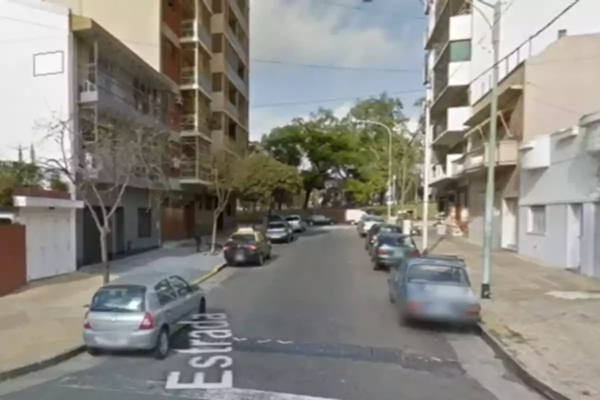 Una calle urbana con edificios altos a ambos lados, varios autos estacionados y árboles al fondo. Una calle urbana con edificios altos a ambos lados, varios autos estacionados y árboles al fondo.