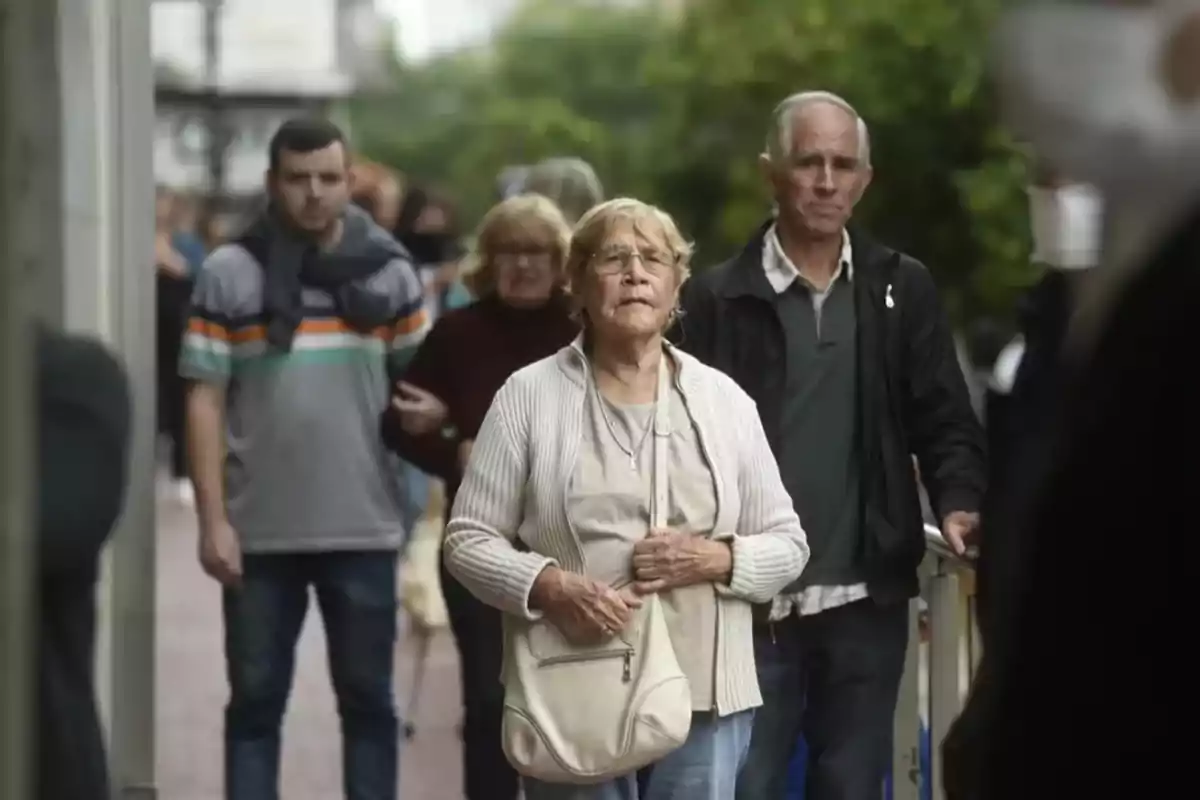 Un grupo de personas camina por una acera en un entorno urbano, con una mujer mayor en primer plano llevando un bolso claro.