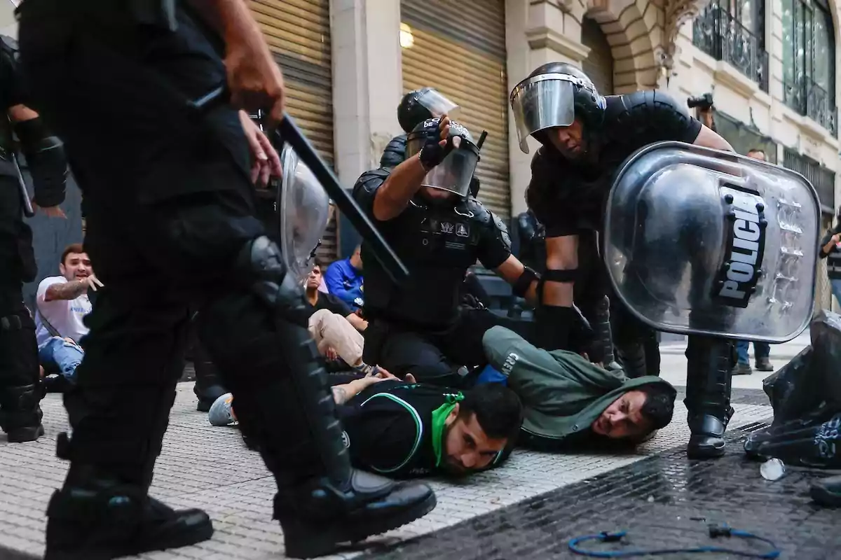 Riot police detain people on the ground during a protest on an urban street.