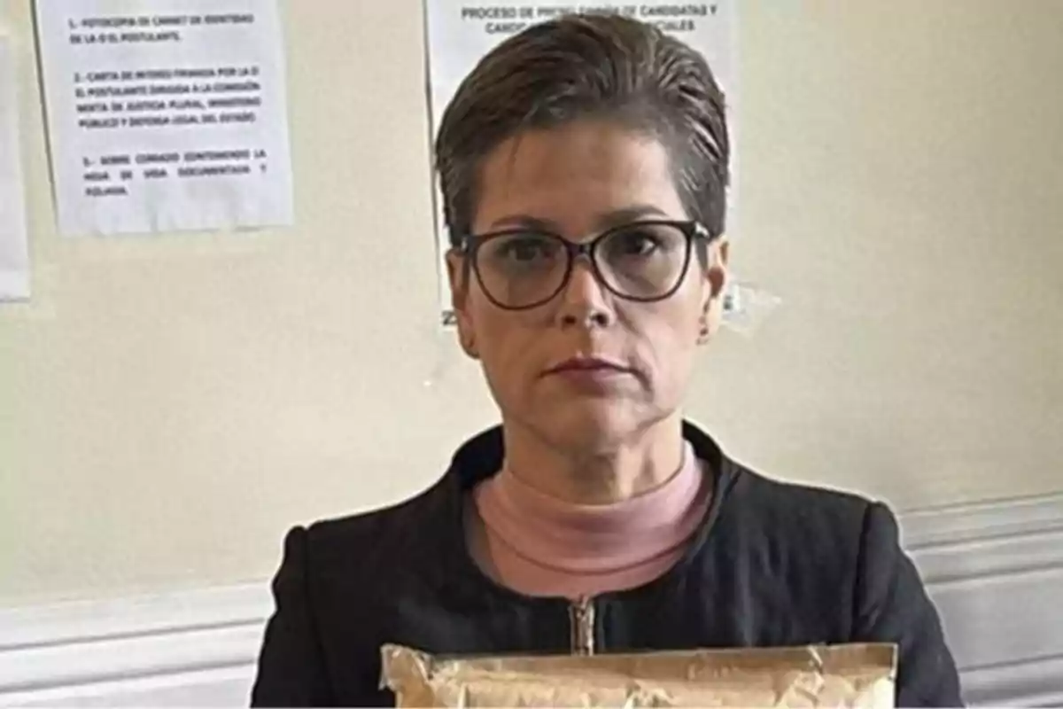 A person with glasses and short hair holds a package in front of a background with papers stuck to the wall. A person with glasses and short hair holds a package in front of a background with papers stuck to the wall.