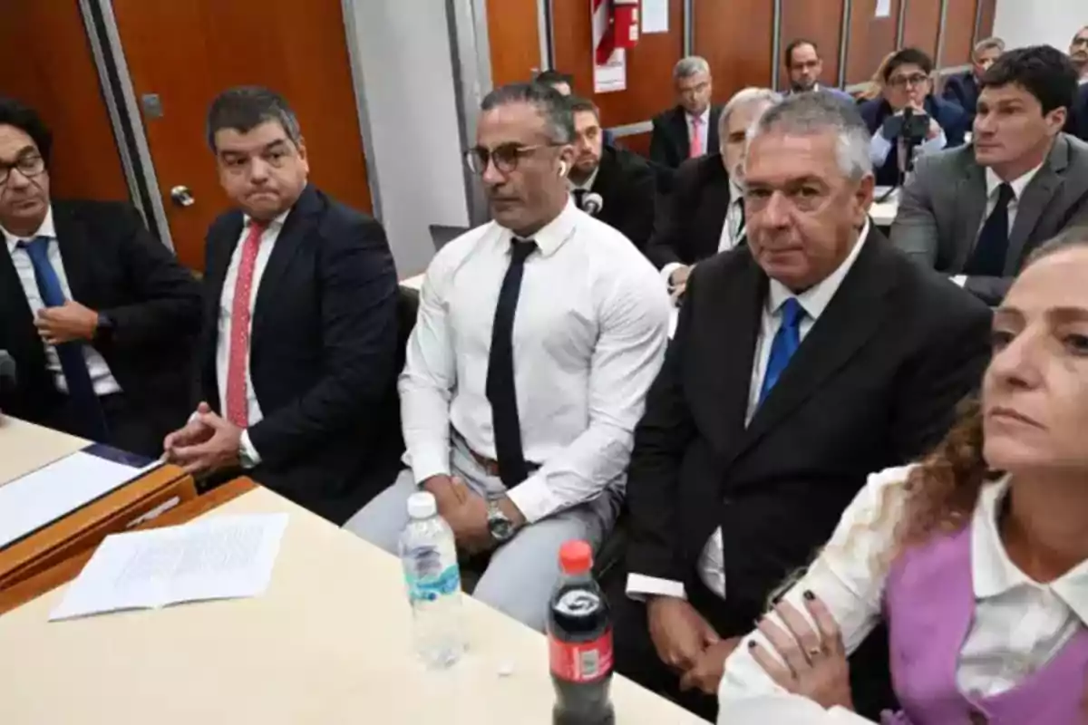 Un grupo de personas vestidas formalmente está sentado en una sala, posiblemente en un entorno de reunión o conferencia, con documentos y botellas de agua sobre la mesa.